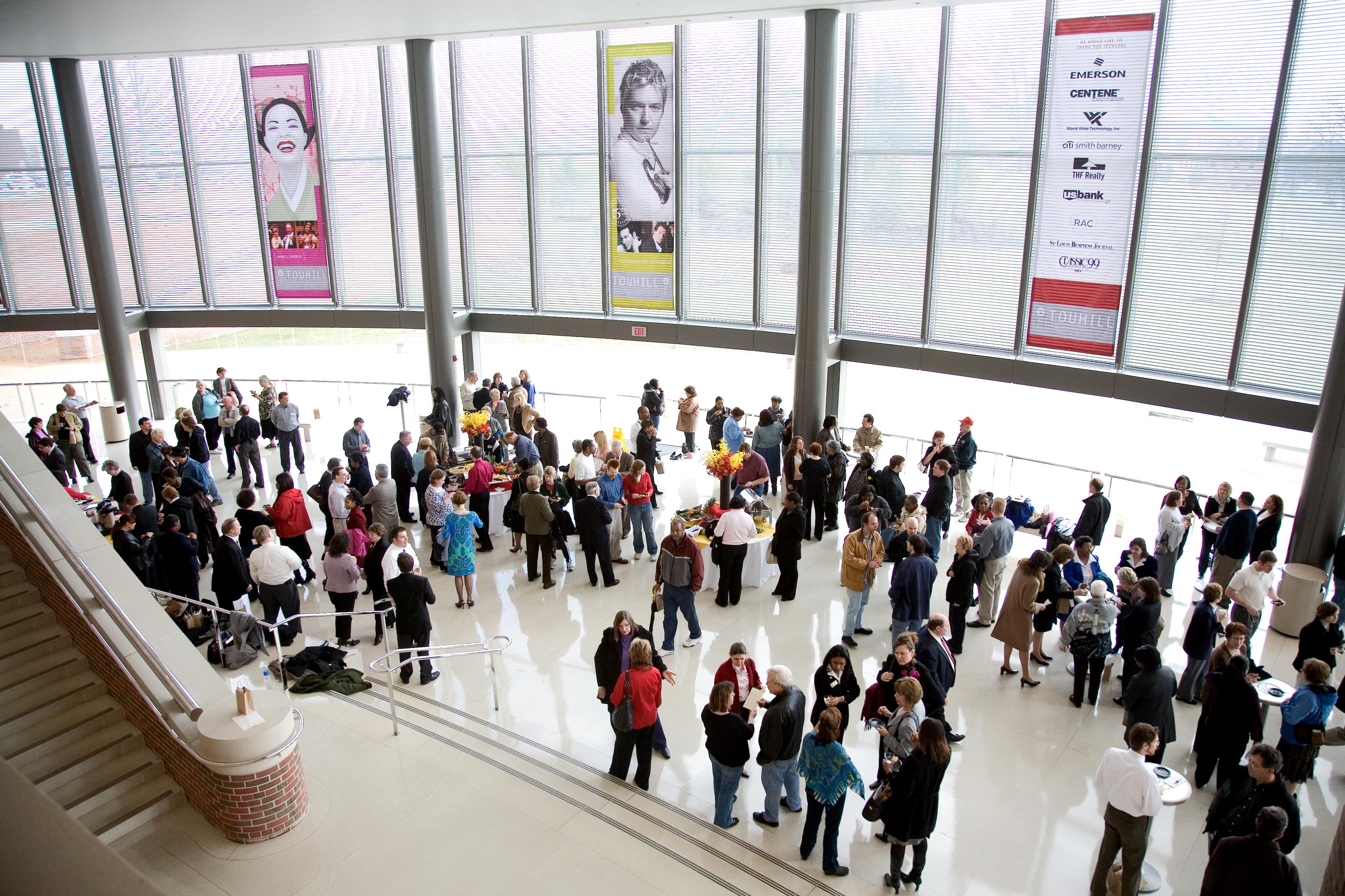 A bustling indoor event with a diverse crowd engaged in conversation, surrounded by large banners and natural light streaming through the windows.